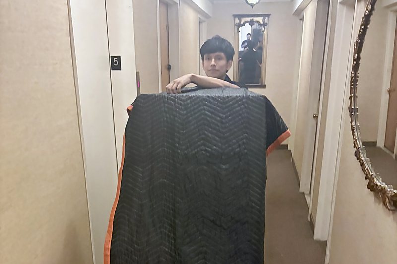 A Move It Clear It crew member carrying a tall item wrapped in a padded moving blanket along an apartment hallway, past the elevator on the fifth floor.